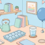 Pastel desk scene with ice cubes, a glass of water, a balloon, a kettle and a jar of pebbles on a blue background.